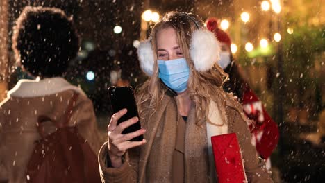 close-up view of happy beautiful young woman wearing earmuffs and facial mask typing on smartphone on the street while it¬¥s snowing in christmas