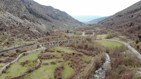 aerial: passenger train in a valley crossing a river on ancient viaduct in the pyrenees, southern france