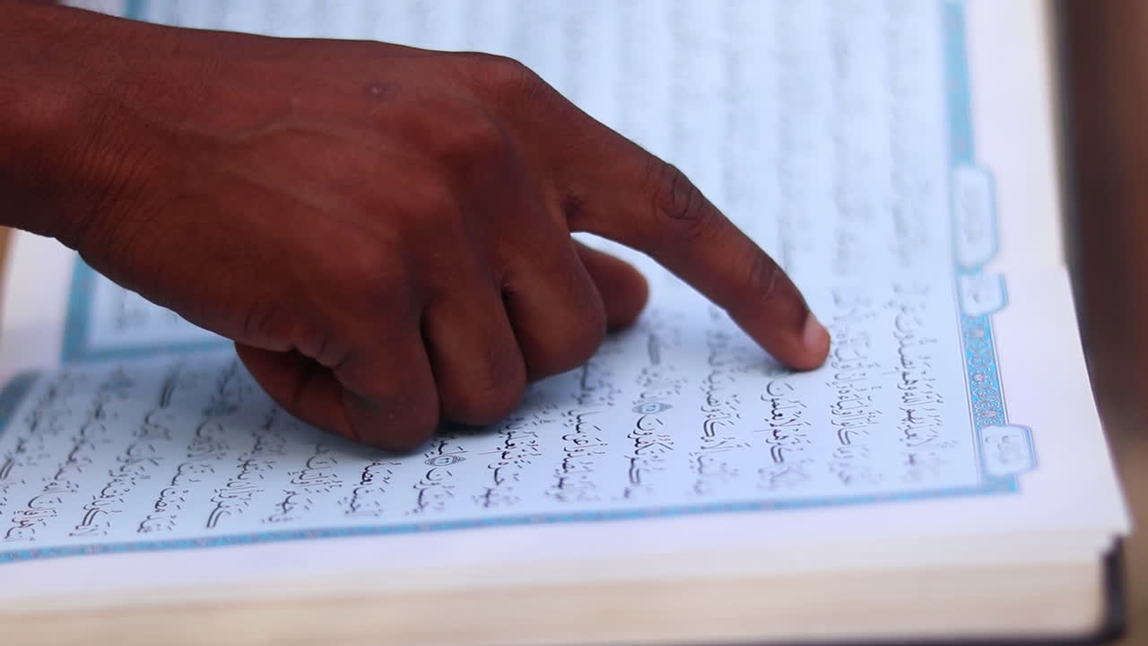 Close up of man using his finger to read the text in the holy Quran while sitting outside on a sunny day