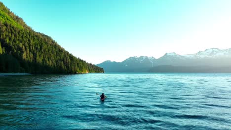 An-aerial-of-a-man-in-a-small-canoe-boat-sailing-in-the-ocean-in-Alaska-with-mountains-in-background