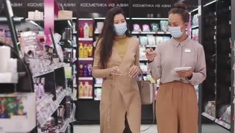 beauty advisor and female customer in masks in make-up shop
