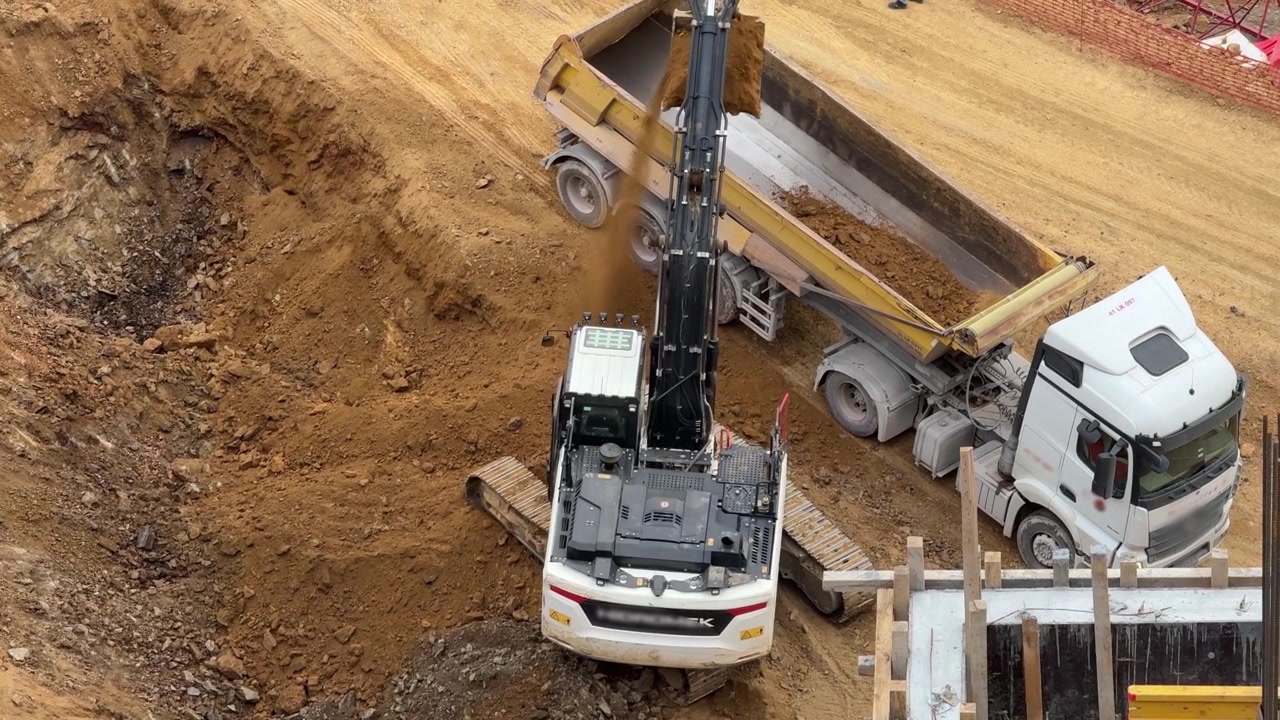 Crawler excavator loading soil and stones into the tipper of a white dump truck at an open construction site. Heavy machinery logistics field operation.