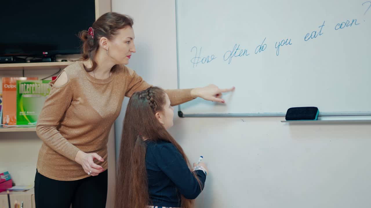 Teacher and little girl in the classroom. Female teacher explains the pupil how to write on board. Clever schoolgirl writing with a marker on magnetic board.