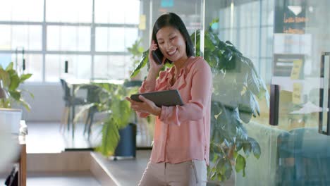 happy asian businesswoman using tablet and talking on smartphone at office, in slow motion