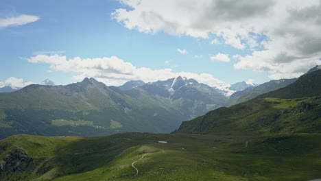 drone flight shows the swiss mountainside with clouds on the blue sky, moving slowly backwards and revealing a small pond with clear blue water on a meadow