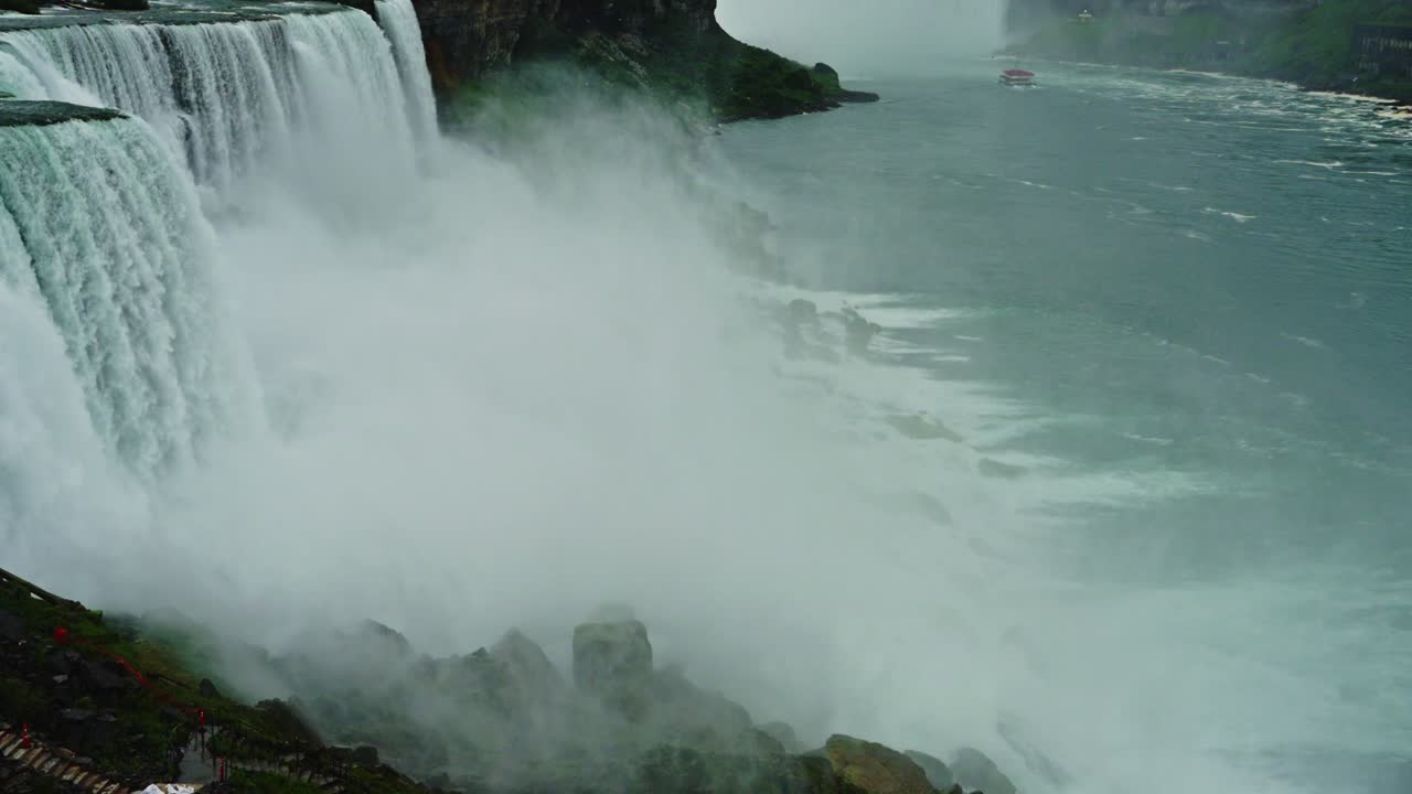 Thick mist rises as water crashes down Niagara Falls, with the river stretching out and a sightseeing boat visible in the distance