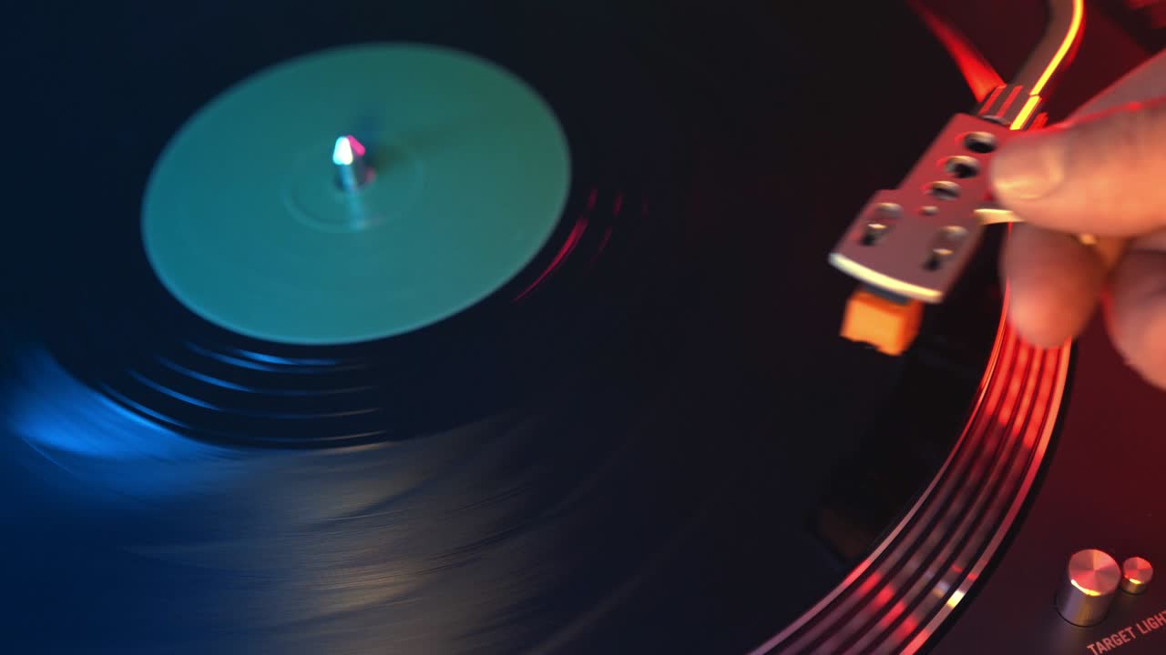 The player vinyl turntable close-up on a background of red and blue lights. The young man's hand smoothly places the needle on the rotating plate. DJ at a youth student's party