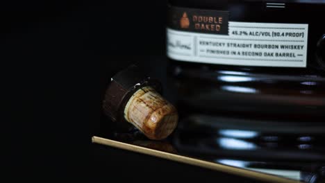 wet wooden cork next to a bottle of double-oaked 90-proof Kentucky straight bourbon whiskey sitting on a mirror with reflections and a dark black background