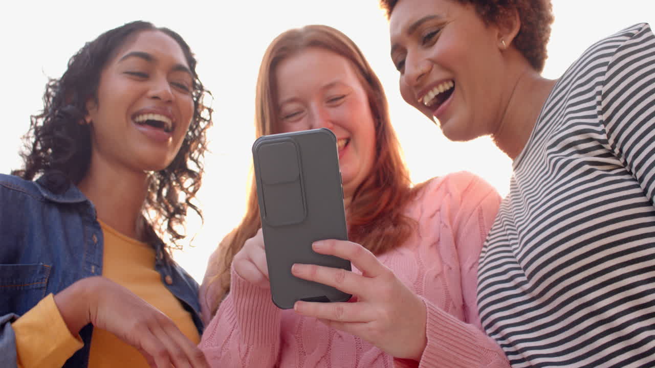 Looking at smartphone, diverse female friends laughing together during hangout