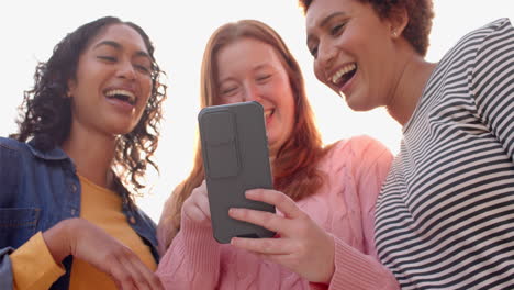 looking at smartphone, diverse female friends laughing together during hangout