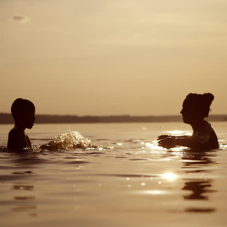 silhouette of a woman and a boy in water at sunset. mother and son are splashing water at each other in the river in the evening.
