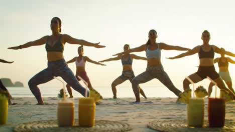 group of women practicing yoga on a beach at sunrise, engaging in warrior pose with refreshing smoothies in the foreground