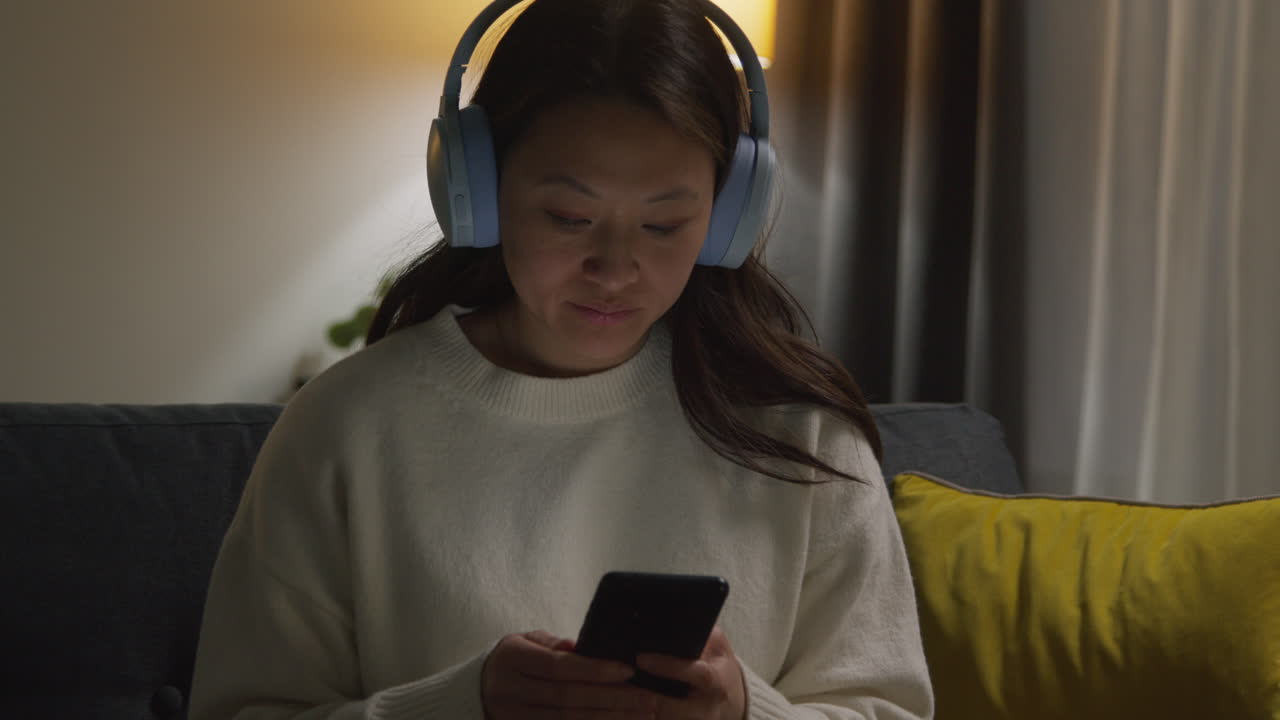 무선 헤드폰을 착용하고 휴대 전화로 음악이나 캐스트를 스트리밍하는 여성이 소파에서 집에서 저녁을 보내고 있습니다.