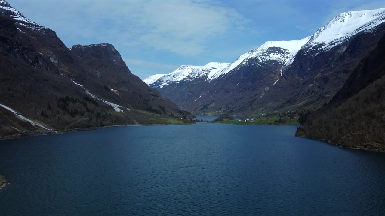 Aerial view of a Norwegian mountain valley with a turquoise lake surrounded by snowy peaks and scattered buildings.