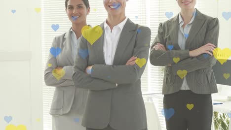 iconas de corazón amarillo y azul sobre tres mujeres de negocios diversas con los brazos cruzados sonriendo en la oficina