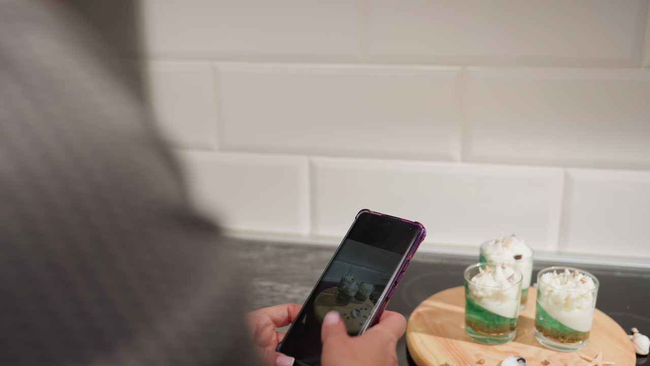 Close up of creative reviewing captured photos on smartphone before switching camera mode to snap more pictures of three beautifully decorated dessert candles placed on wooden tray in kitchen setup