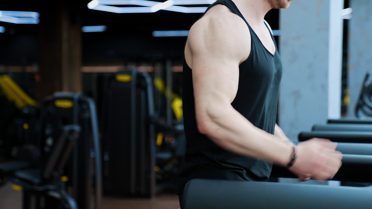 Sportsman on a treadmill. Athletic man with muscular body training on sports equipment in a gym. Fitness guy on a modern simulator. Side view. Slow motion.