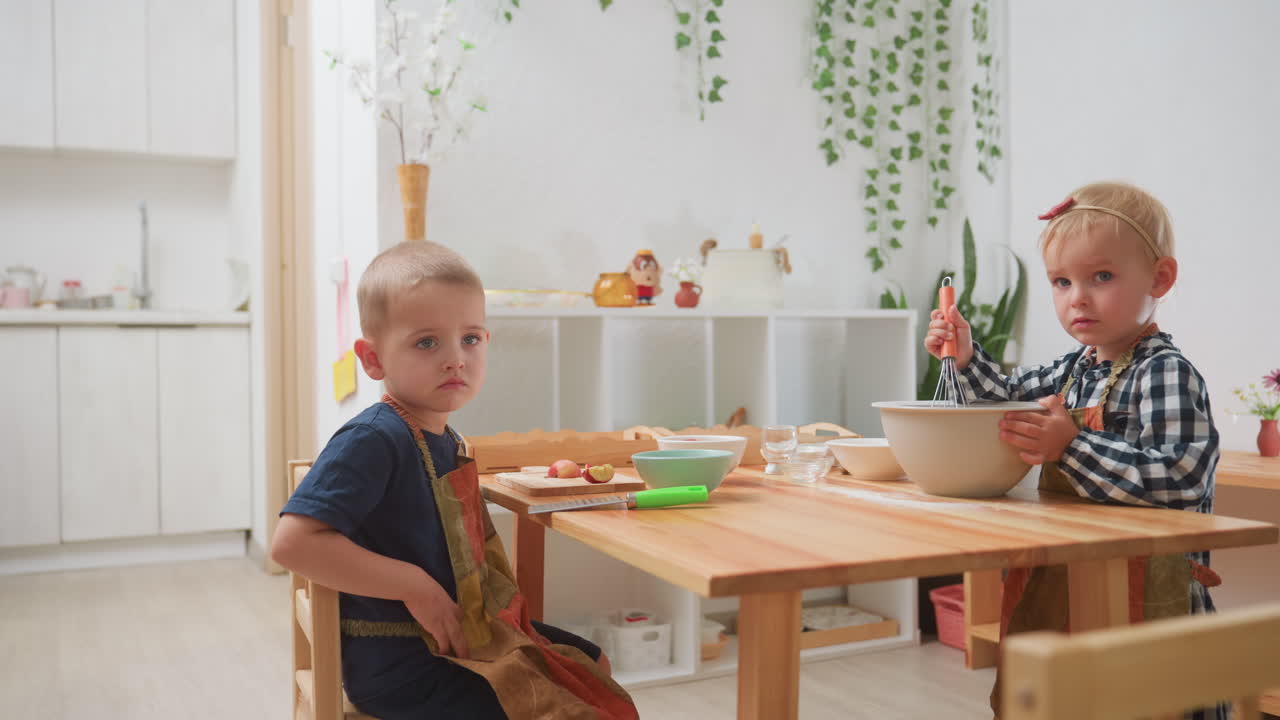 Bambini in un'aula di cucina, un ragazzo seduto stanco con una maglietta blu, una ragazza che mescola l'impasto in una ciotola, un altro bambino con una maglietta a righe che si allontana, ambiente decorato con piante e utensili per la cottura