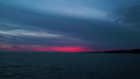 Drone-pull-back-from-pink-red-glow-below-dark-grey-blue-clouds-above-water