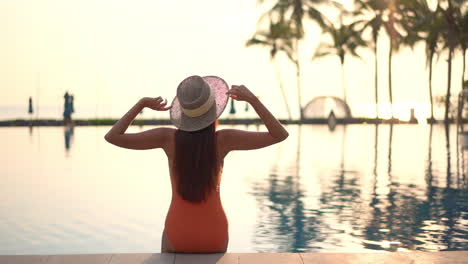 a woman with her back to the camera raises her hands into the air greeting the sun rising over a swimming pool and the ocean horizon beyond