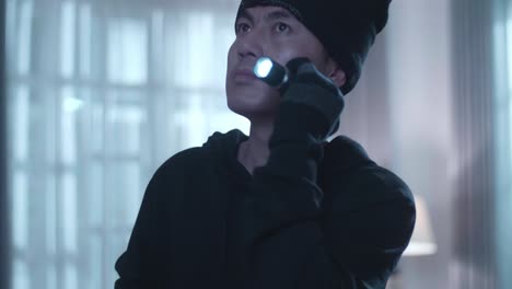 close up of asian thief man holding flashlight looking around and putting a mask down while walking in someone's house