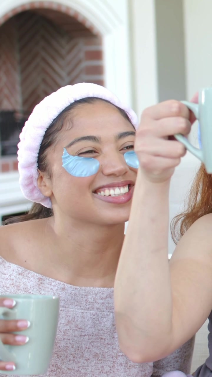 Vertical video: Smiling friends in pink and gray clinking mint mugs on sofa, wearing blue patches