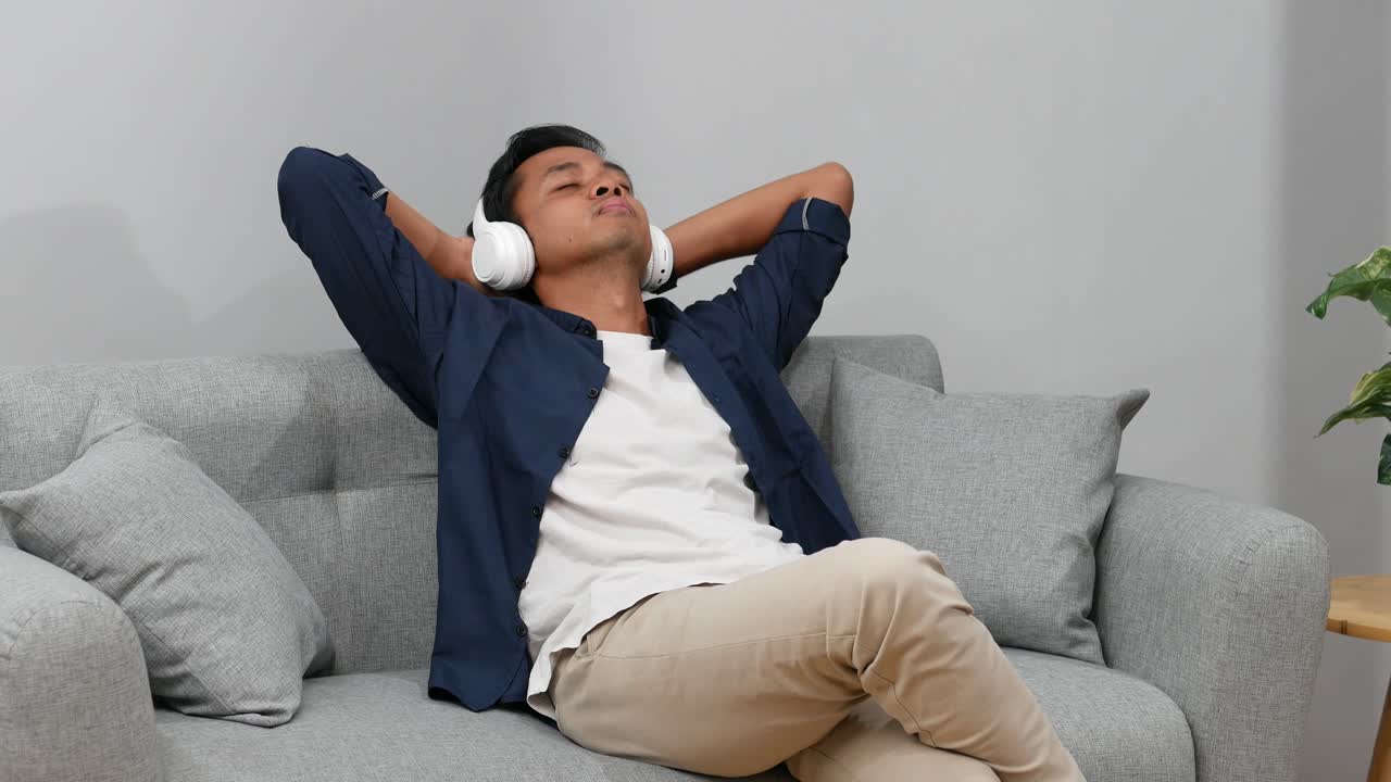 Giovane uomo asiatico che trascorre del tempo di qualità seduto sul divano di casa, ascoltando musica con le cuffie.