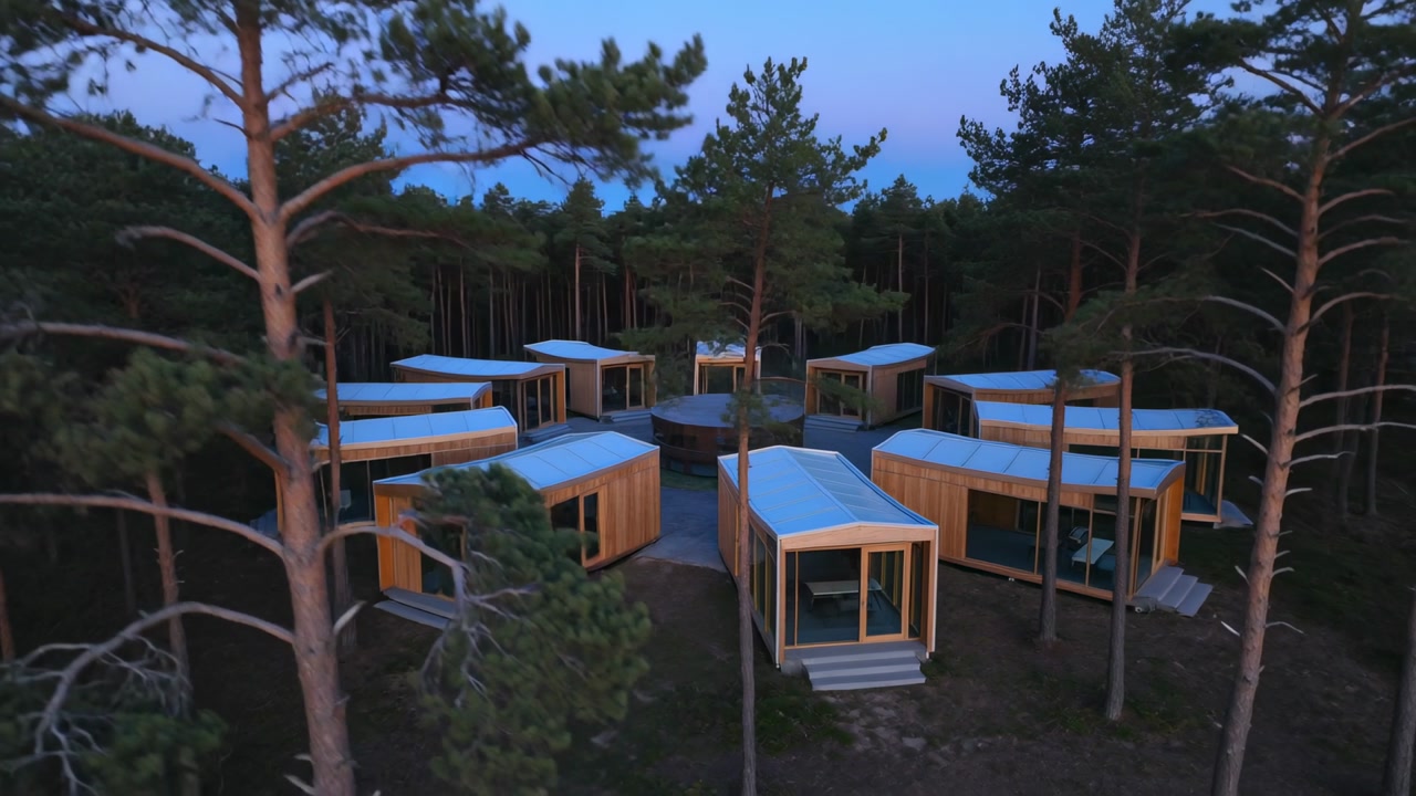 Approaching drone capturing wooden cabin cluster in pine clearing, showing glass facades, decks
