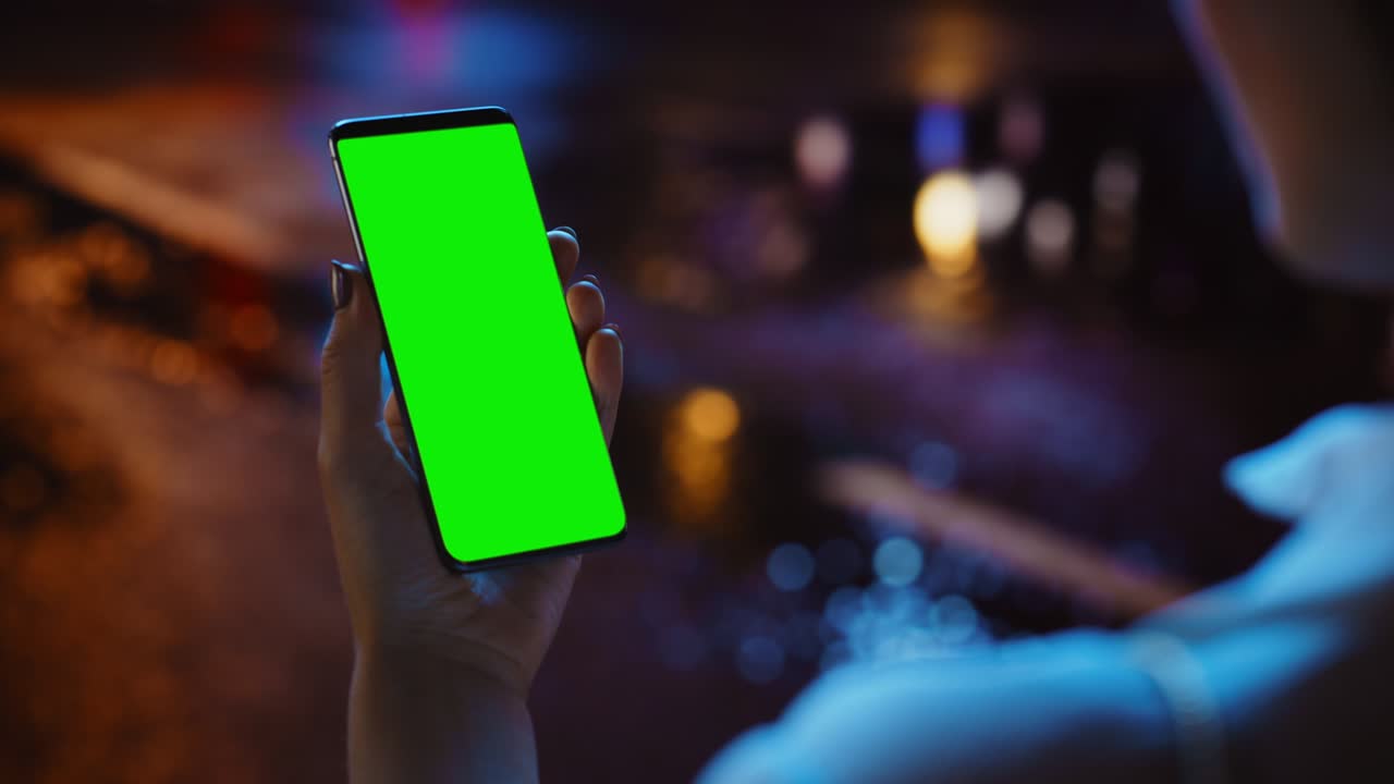 Woman using Chroma Key Smartphone while Standing, Looking at the Green Screen. Night City Street Full of Neon Light. Female Using Mobile Phone. Over the Shoulder Closeup Tracking Shot Focus on Display