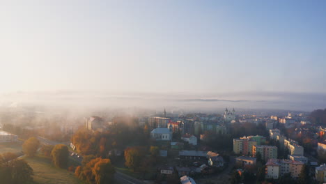 drone shot of a mist covered town at sunrise, bathed in soft morning sun. light illuminates the fog creating a warm, vivid, unique, autumn landscape. blue sky adds color contrast. sanok, poland