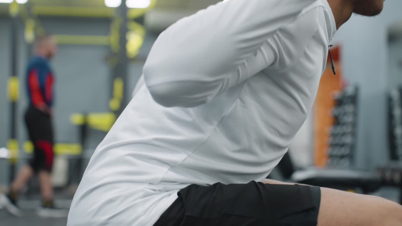 side view of student squatting with barbell under pressure in modern gym environment while other people train in background, wearing black shorts with reflective patch on leg