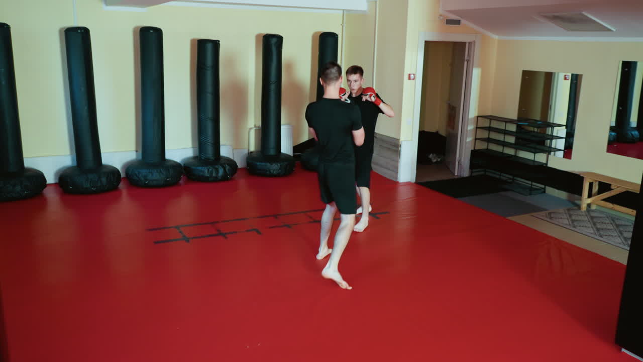 Wrestlers sparring inside gym, wearing gloves and black outfits, practicing combat sport movements with defensive stance and striking preparation on red mat floor surrounded by standing punching bags