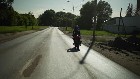 back view of a female motorcyclist standing as she rides a power bike close to a muddy path, empty road, car approaches in the opposite direction, with trees and buildings in the background