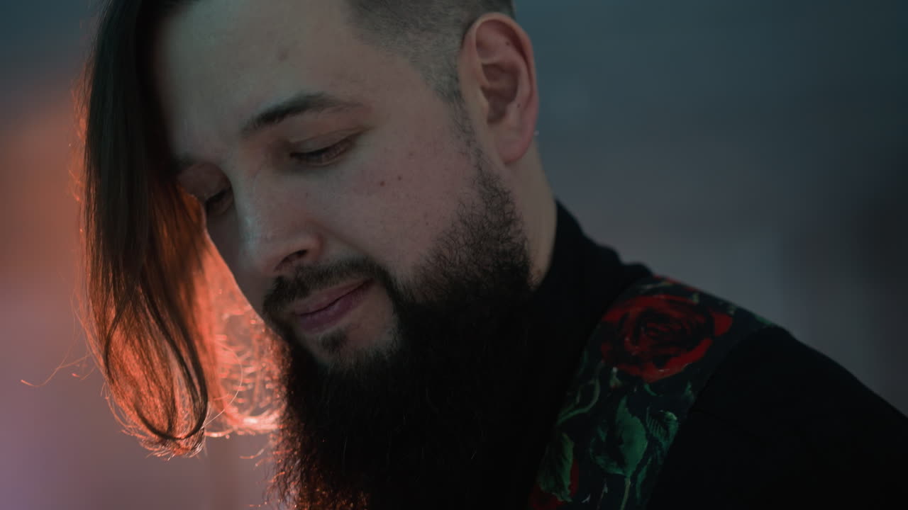 бородатый гитарист в черной рубашке играет на гитаре в мягко освещенной, размытой комнате. окружающее освещение подчеркивает сосредоточенное выражение лица музыканта