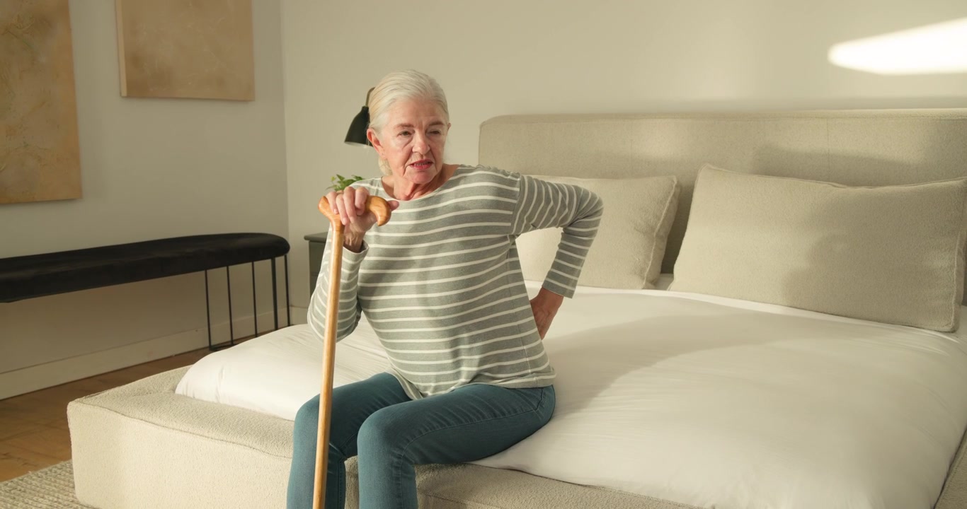 Donna anziana con maglia a righe seduta sul letto, che tiene un bastone di legno, alleviando il mal di schiena e aggiustandosi.