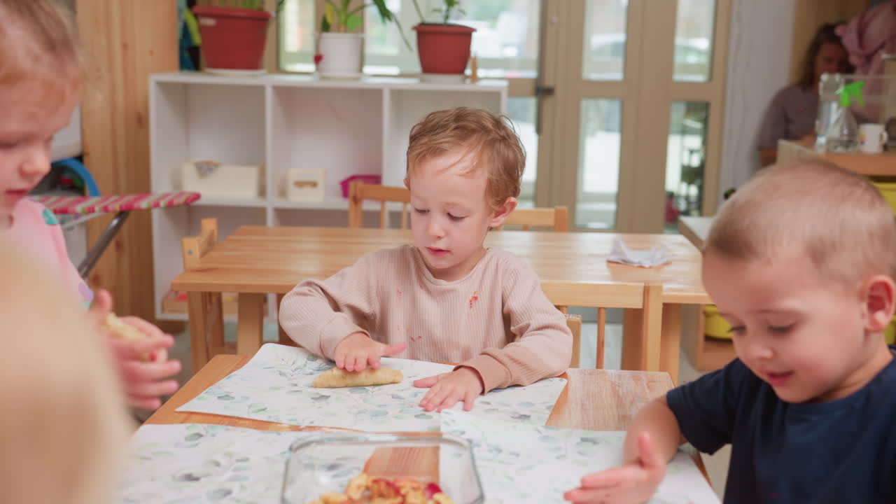 Kindergartenkinder rollen Teig auf dem Tisch während einer Unterrichtsaktivität, die sich auf Kreativität, Geduld und Kompetenzentwicklung konzentriert, während junge Lernende Texturen und Formen erkunden
