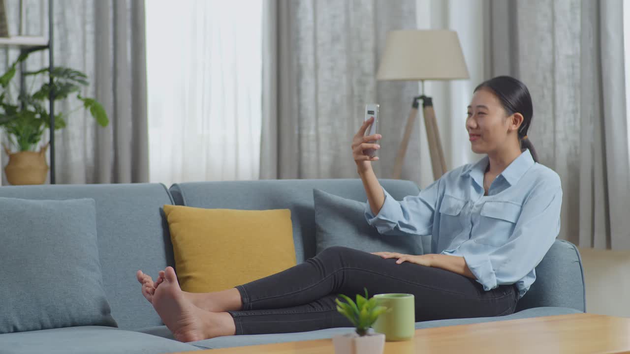 아시안 여성의 몸이 가득 차 있고, 손을 흔들며 거실의 소파에 누워있는 동안 스마트폰으로 비디오 통화를 하고 있습니다.