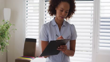 Portrait-of-mixed-race-female-physiotherapist-holding-clipboard-and-writing-looking-at-camera