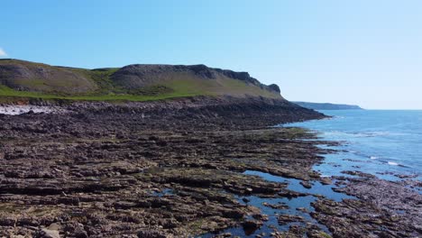 sideways drone view of rocky coastline in gower with rugged cliffs and small rock pools with land in far distance creating parallax effect.