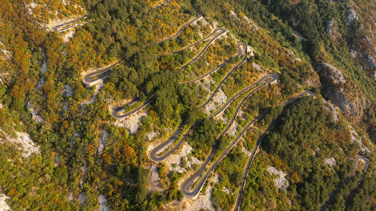 Tornanti a gomito serpeggianti si snodano su un ripido versante montuoso di Cattaro, ricoperto di alberi, sotto una calda luce diurna, visti da un sorvolo di drone ad alta quota