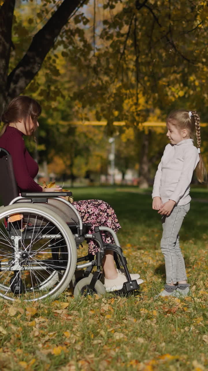 madre e hija se divierten en el parque en un soleado y cálido día de otoño. dama en silla de ruedas sostiene hojas secas y se comunica con niña preescolar sonriendo felizmente