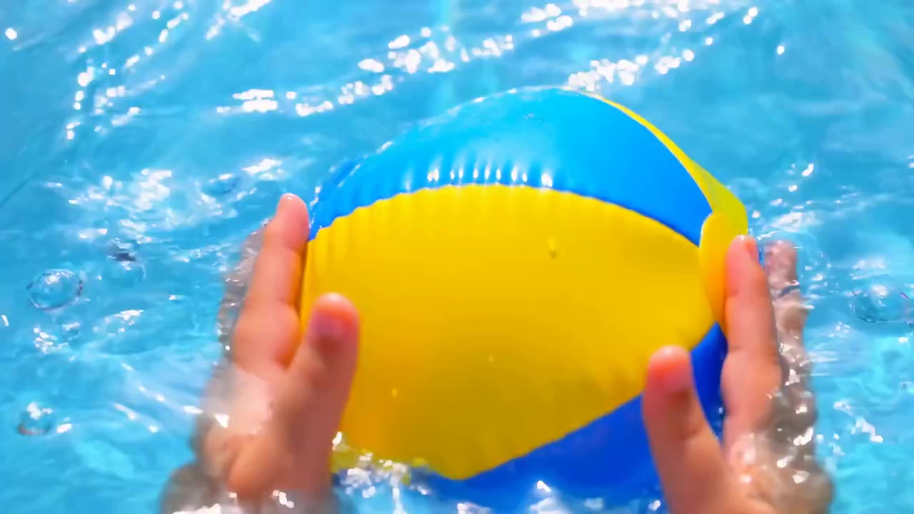 Hände halten und stabilisieren einen blau-gelben Wasserball auf der helltürkisen Pooloberfläche, während Wasser um die Handflächen schwappt und Tropfen spritzen, was lebendige Sommerfarben, Schimmer und spielerische Outdoor-Energie erzeugt