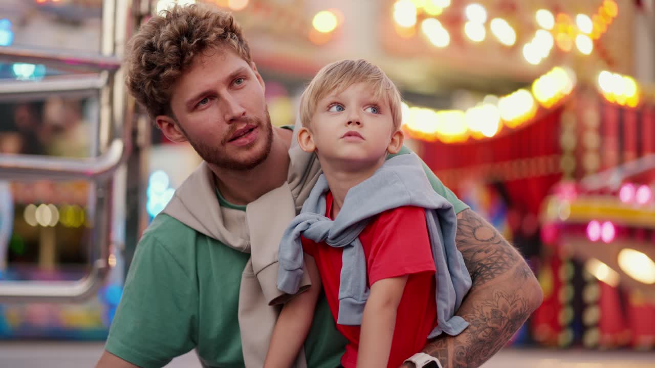 빨간 티셔츠를 입은 작은 금발 소년은 아버지의 다리에 앉아 놀이 공원의 밝고 다채로운 명소들을 함께 보고 소통합니다.