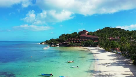 Fishing-boats-floating-on-shallow-turquoise-lagoon-near-white-sandy-beach-palm-trees-and-holiday-resort-in-Thailand