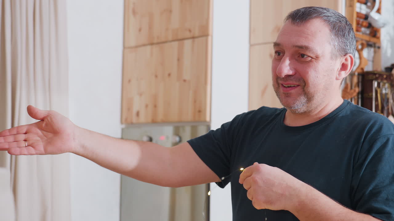 Man in green top smiling while holding Christmas light indoors preparing festive decorations with joyful expression surrounded by cozy warm atmosphere of holiday season and cheerful family spirit