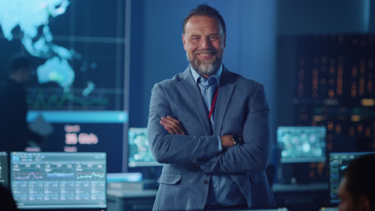 leader di progetto maschio senior di successo in un ufficio di ingegnere informatico in piedi con le braccia incrociate. guarda la telecamera e sorride. controlla la stanza di monitoraggio con una mappa globale su un grande schermo