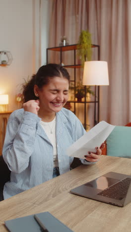 excited young indian woman freelancer sitting at office desk opening envelop letter and reading it