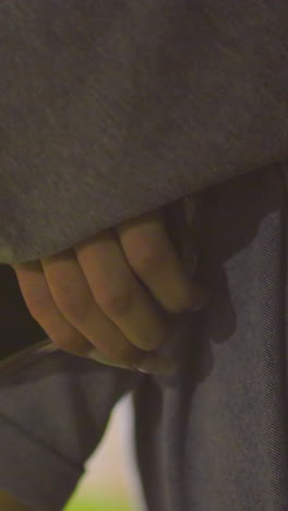 primer plano de un individuo con un teléfono móvil en la mano, con una manga gris y vaqueros, con una iluminación de fondo suave y un paisaje borroso del parque que crea una atmósfera tranquila por la noche