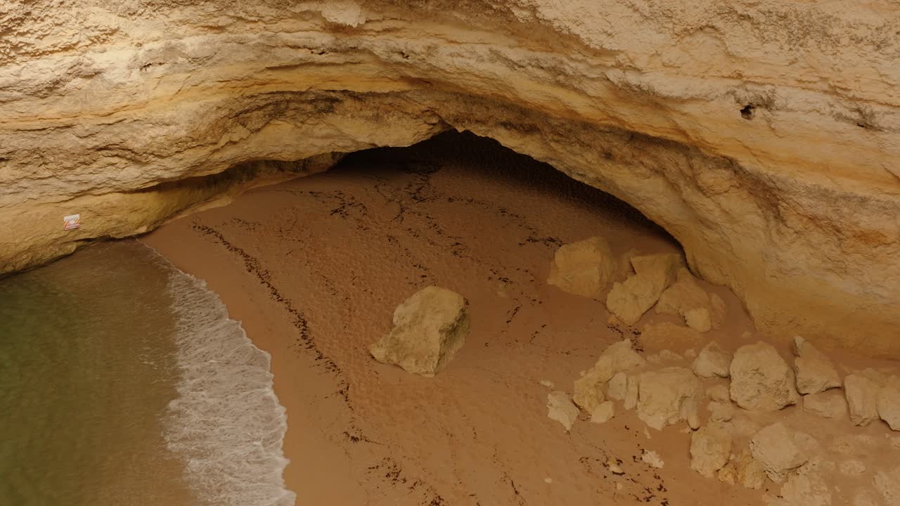 Close aerial view of a secluded sea cave with sandy beach and fallen rocks, captured during low tide in Algarve, Portugal