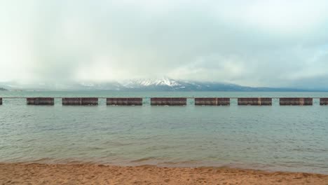 South-Lake-Tahoe-Durante-El-Invierno-Con-Una-Playa-De-Arena-Dorada-En-Primer-Plano,-Un-Muelle-En-El-Medio-Y-Magníficas-Montañas-Nevadas-En-El-Fondo---Plano-Amplio